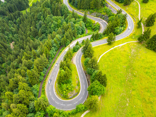 Naklejka premium Curved asphalt paths wind through lush green forests in the Alps, offering breathtaking views and a thrilling drive. This aerial perspective captures the beauty of a summer road trip.