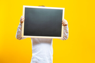 Preschooler Caucasian Boy Holding Wooden School Blackboard Pointing Forward With Lifted Hands Hiding Face Isolated On Yellow Background as Image To Portray School Preparation Ideas