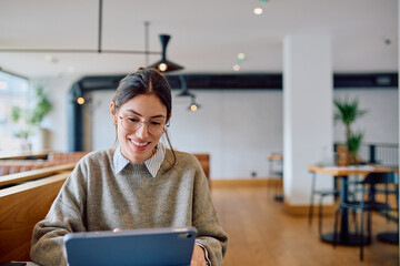 Young woman wearing glasses smiling while working or studying on a digital tablet in a bright, contemporary cafe