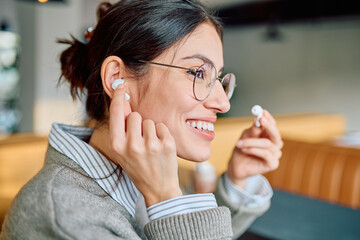 Young woman smiling, wearing glasses, inserting wireless earbuds into her ear, enjoying audio...
