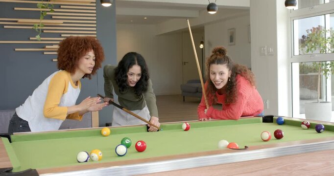 Three female friends holding cues around pool table in game room guiding shot celebrating victory