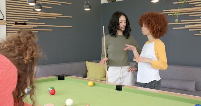 Female friends discussing angles at pool table, urging third striking cue pocketing ball, cheering