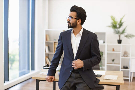 An Indian businessman in a suit stands in a modern office, looking out the window - Powered by Adobe
