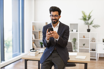 An Indian businessman in a suit smiles while using his smartphone in a modern office setting