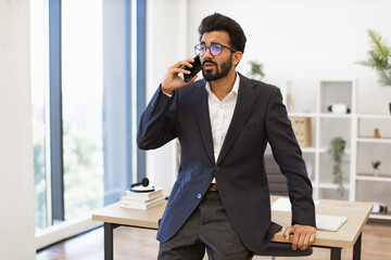 An Indian businessman wearing glasses is talking on the phone in his office, looking concerned