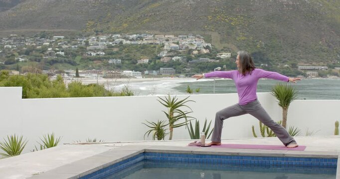 Mature woman stepping barefoot on pink mat poolside, pivoting into hip-opening straddle pose