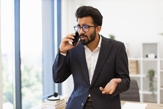 An Indian businessman wearing glasses looks concerned while talking on his phone in his office