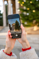 Close-up of hands holding a smartphone while photographing the Temple of Augustus and a decorated Christmas tree in Pula, Croatia. Festive holiday atmosphere with warm bokeh lights