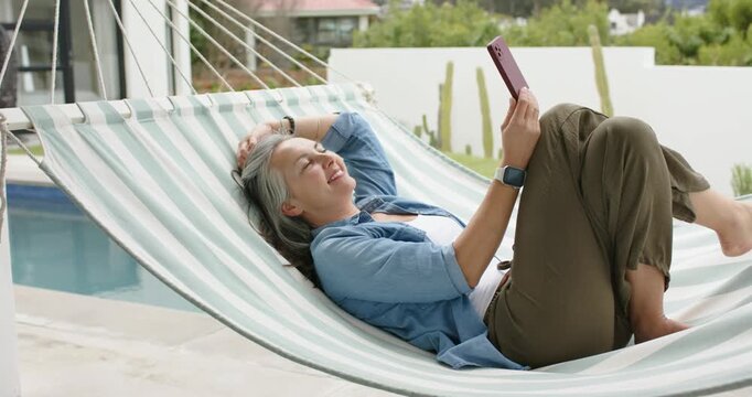 Senior woman reclining in hammock by pool scrolling pink tablet while brushing hair and glancing