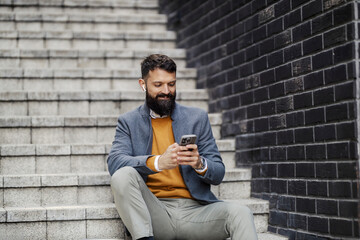 Portrait of smart casual businessman sitting on staircase with cellphone in hands at financial district and texting messages,