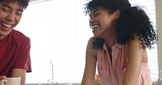 Diverse couple touching hands by mugs, laughing and sipping coffee, connecting at kitchen counter