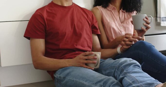 Couple sitting on kitchen floor holding mugs, linking fingers, man resting head on woman's shoulder