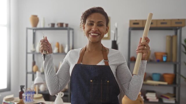 Woman holds pottery tools and a wooden dowel in both hands in artisan studio with shelves of clay pots and tools; creative pride.