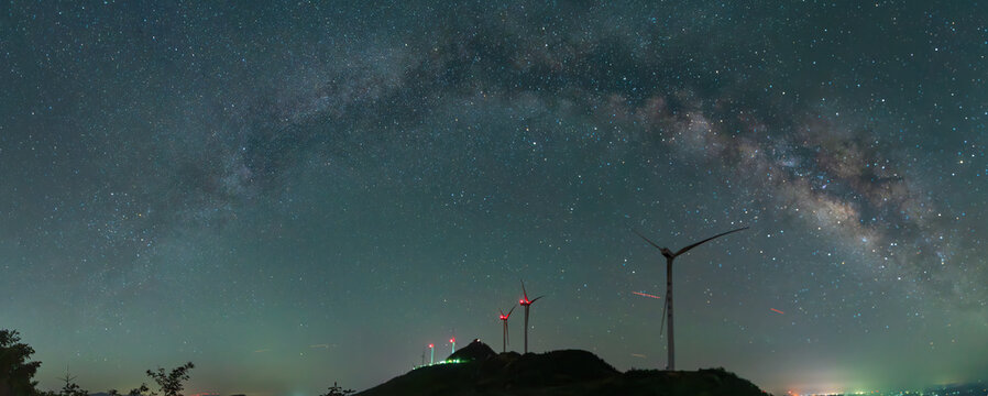 Silhouette of wind turbines under a starry night sky with the milky way visible, hills and trees in the background at dusk