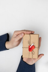 Two hands are holding a small, wrapped gift. The gift is adorned with twine and a red heart clip. It is perfect for celebrating Saint Valentine's Day.