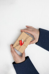 Two hands hold a small gift box tied with twine and a red heart decoration. The setting is simple and suggests a moment of giving for Valentine's Day.