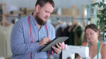 Young bearded tailor in checked shirt sitting on edge of worktable, sketching clothing designs on tablet with focused, creative look while female colleague working at sewing machine in cozy workshop