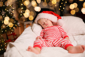 Baby in Christmas pajamas and Santa hat sleeping on bed indoors