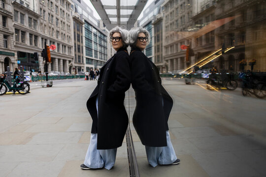 Fashionable woman with glasses standing against reflective wall in city