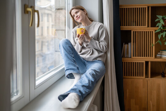 Happy dreamy woman enjoys quiet time pause holding coffee mug relaxing sits on windowsill. Serene smiling female takes break feeling inner peace, self-reflection tranquil mood. Gratitude, contentment
