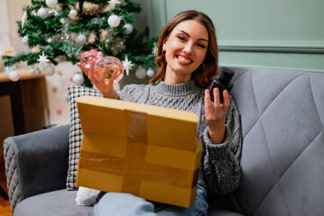 Happy woman sits comfortably on a sofa, holding perfumes and unboxed delivered package. Decorated Christmas tree in the background.