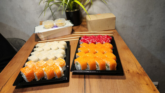 Sushi set in black tray placed on wooden table with green houseplant in background, grey wall, closeup photo for online food delivery, sushi restaurant menu, catalogue, or culinary website content. - Powered by Adobe