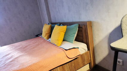 Double bed with orange blanket, yellow and blue pillows, grey wall, bar stool by headboard, minimal modern bedroom in apartment, simple setup, photo for real estate listings, rental apartment websites