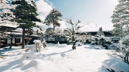 A serene traditional Japanese garden blanketed in snow, featuring pruned trees and subtle sunlight filtering through the branches