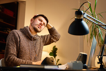 Man sitting at desk stretching neck by tilting head with hand while working on laptop. Concept of office work, computer fatigue, muscle tension and stress relief exercises