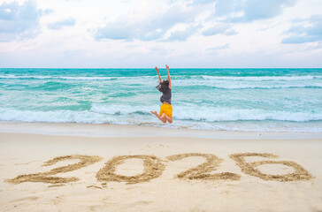 New Year 2026 written on sand, happy woman with hands up jumping on the beach