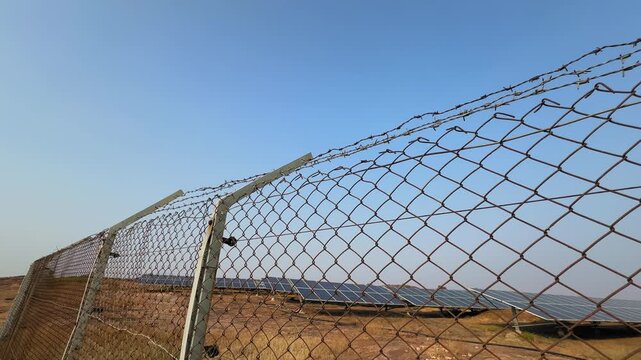 Panning view of a vast solar farm behind a chain-link fence under a clear blue sky, showing long rows of solar panels stretching across dry open land.