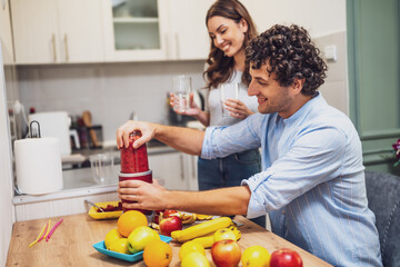 Smiling and relaxed, the two share a moment together while preparing homemade fruit drinks, with a countertop full of oranges, apples, and bananas.