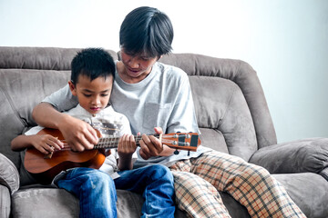 Two Young Asian Brothers Sitting Together On Grey Sofa And Playing Ukulele Together