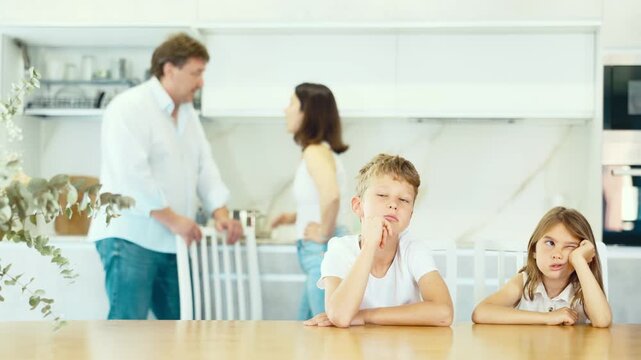 Wrathful mother and father brawling behind their two children sitting sadly at the table in the kitchen
