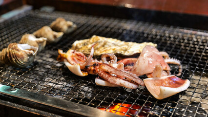 Fresh squid tentacles and whole sea snails are grilling on a dark metal grate over hot charcoal, showing Japanese seafood barbecue