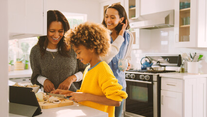 Female Multi Generation Family Prepare Meal In Kitchen Following Recipe On Digital Tablet Together