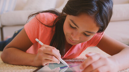 Close Up Of Young Girl Lying On Floor in Lounge At Home Drawing With Stylus On Digital Tablet