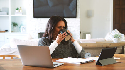 Obraz premium Mature Woman With Hot Drink Sitting At Table At Home Checking Finances On Laptop