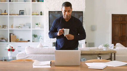 Mature Man With Hot Drink Standing At Table At Home Reviewing Finances On Laptop