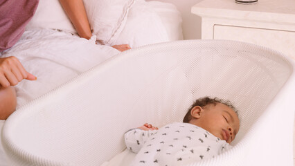 Close Up Of Mother Sitting On Bed Next To Sleeping Baby Son Lying In Cot In Bedroom