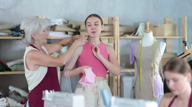 Skilled seamstress with a needle on arm sews fabric against the background of a mature atelier worker who shows a sample to a client. High quality 4k footage