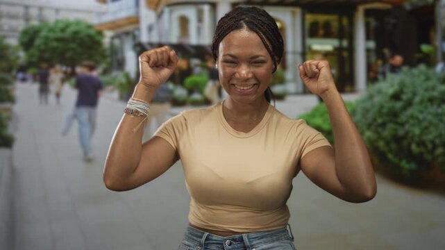 Young woman cheering with fists raised, bare forearms visible, standing on a busy urban street lined with shops; excitement celebration victory triumph.