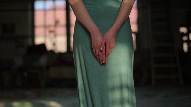 Medium shot of a modest woman in a long, teal-colored dress holding her hands together, standing in a dimly lit, rustic industrial or artist's studio setting with soft natural light.