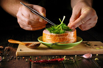 A chef uses tweezers to place fresh herbs on a plate with meat on a wooden surface. Spices and garlic surround the plate, adding to the kitchen atmosphere in the late evening