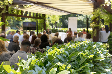 Outdoor business conference with professional audience attending presentation in green garden venue under canopy, sustainable corporate event setting
