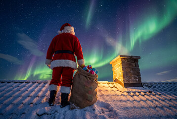 Santa stands on a snow-covered roof next to a chimney and his sack of gifts, looking out at the dramatic green and purple bands of the Aurora Borealis in the night sky.