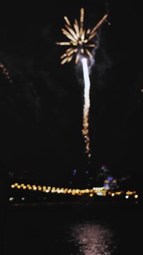 Vertical video. Fireworks explode against the background of a night city by the river. Vertical video. Festive fireworks reflected in the water. Celebration of the city day.