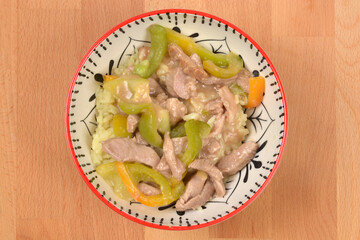 An overhead studio shot of a bowl of pork strips and green bell pepper stir-fry with a creamy sauce served over rice on a wooden background