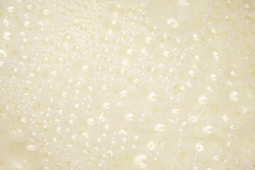 A high-angle, full-frame background of white liquid milk covered in a dense layer of small, frothy, reflective air bubbles