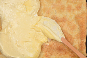 A close-up, high-angle view of a wooden spoon spreading creamy yellow custard filling over a sheet of baked puff pastry on a dark baking tray